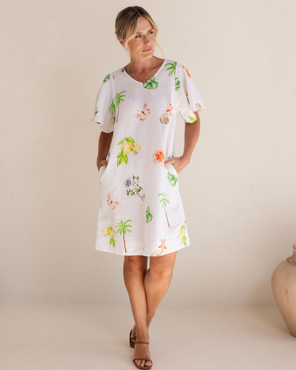 Woman wearing a white dress with tropical print against a plain background. See Saw Linen V Neck Flutter Sleeve Midi Dress