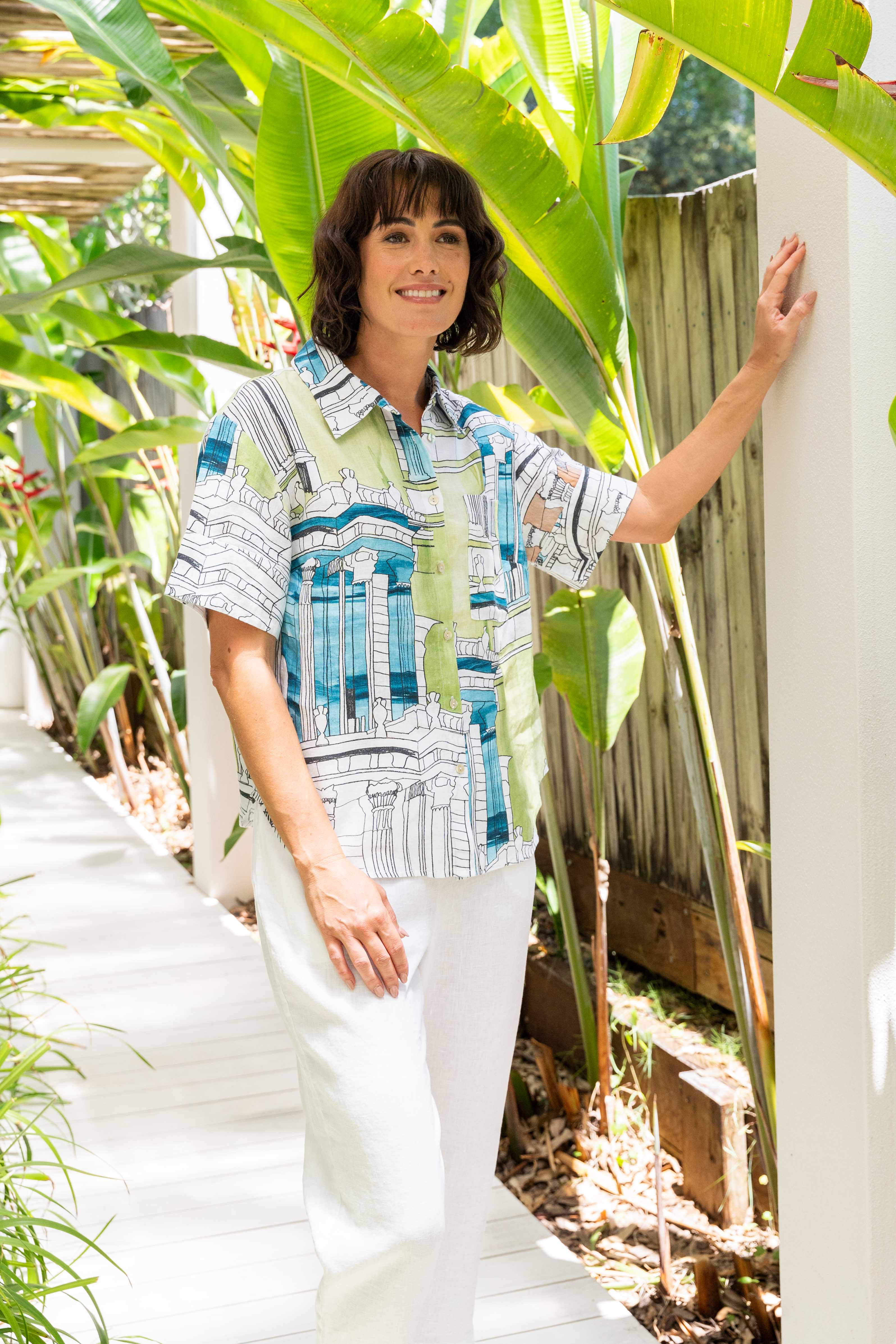 Woman in a patterned shirt and white pants standing in a garden with greenery. See Saw Linen Short Sleeve Shirt