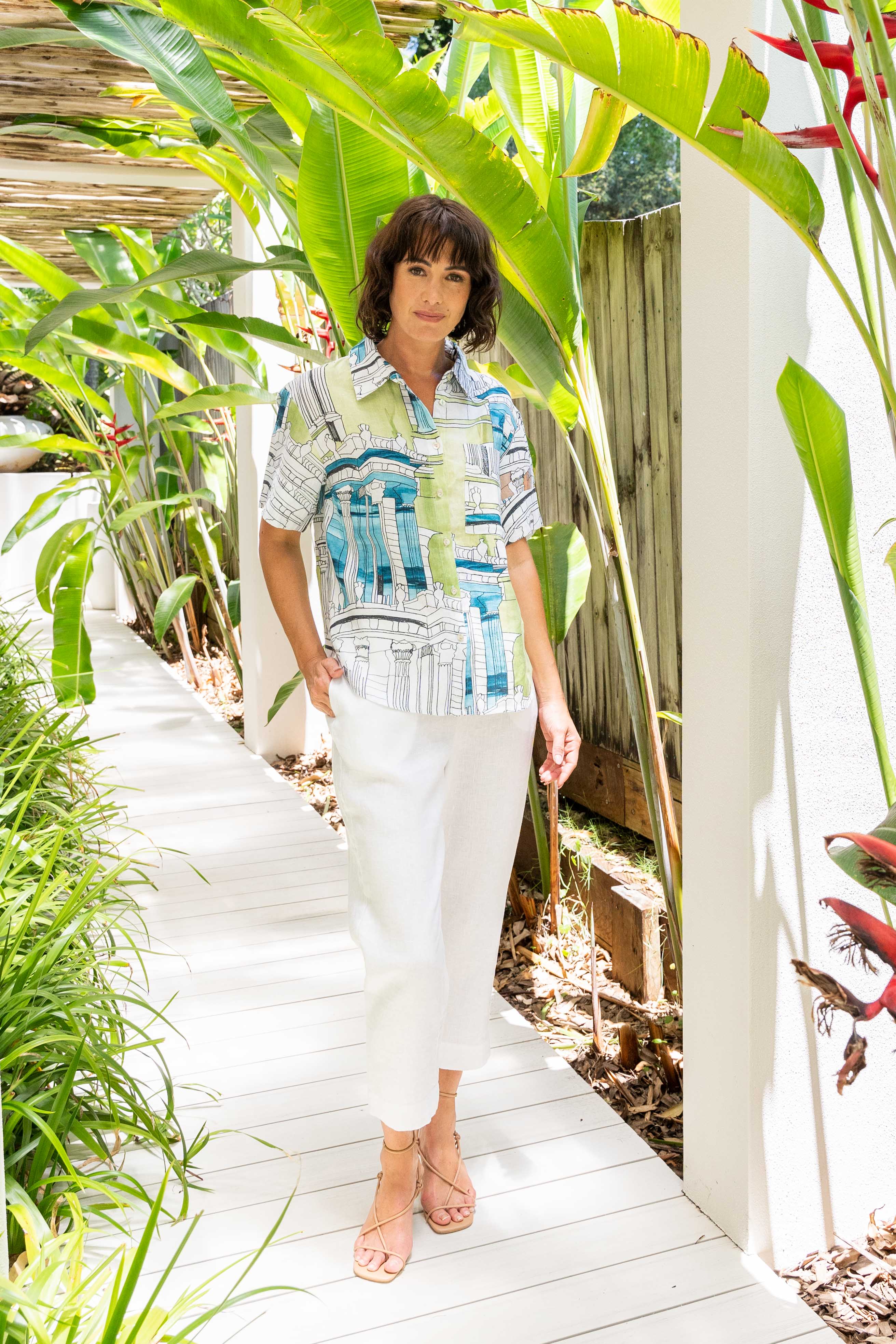 Woman standing in a tropical setting with lush greenery and a white pathway. See Saw Linen Short Sleeve Shirt
