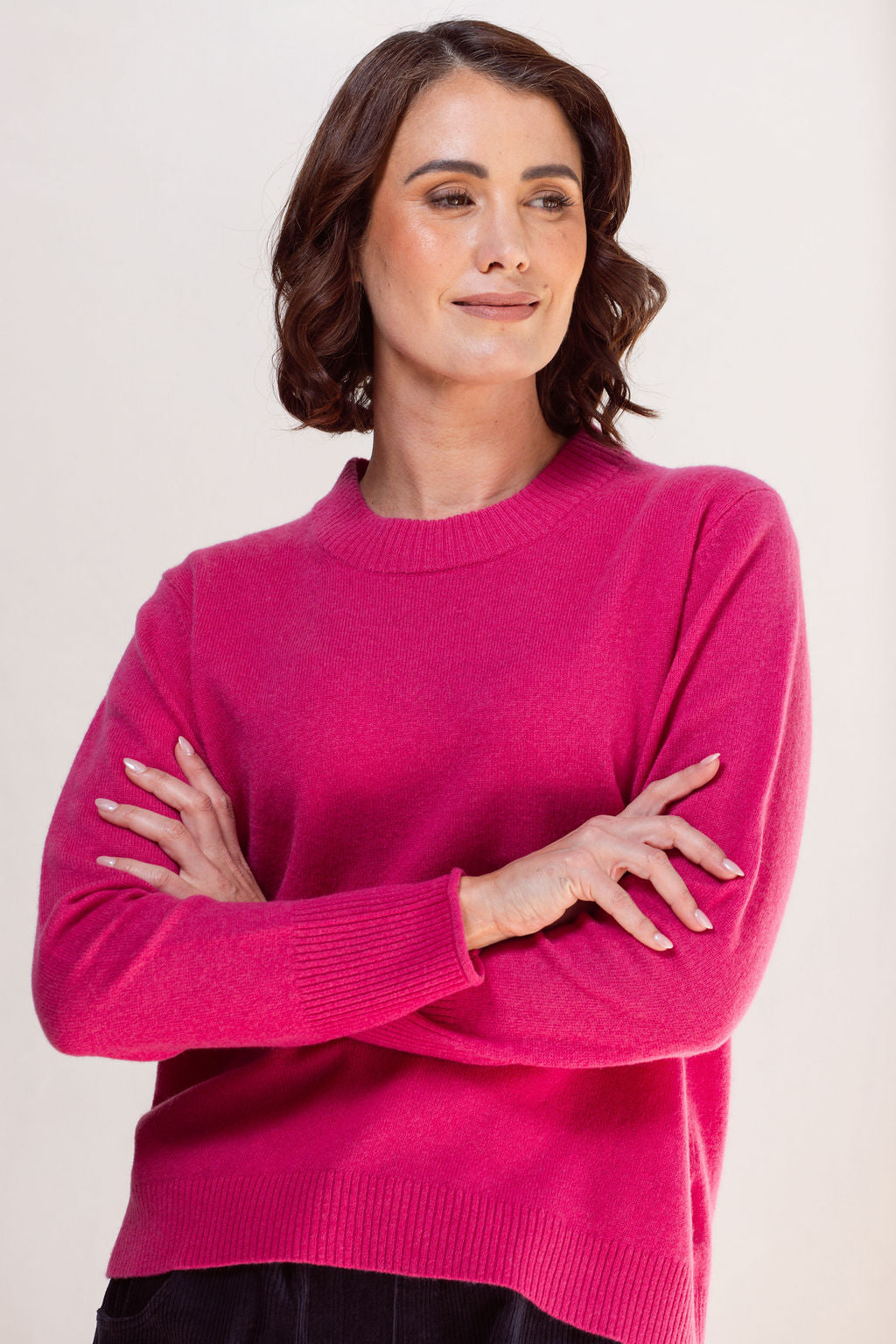 Woman wearing a pink crew neck See Saw sweater and light blue jeans against a plain background