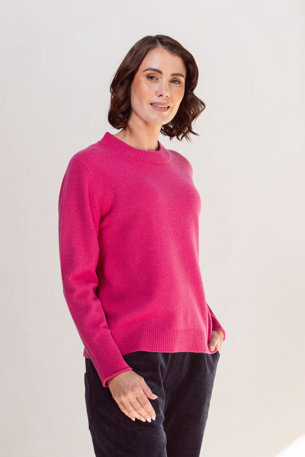 Woman wearing a pink crew neck See Saw sweater and light blue jeans against a plain background