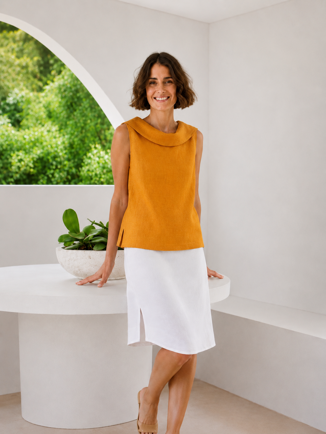 A woman modeling a sleeveless marigold yellow See Saw Linen cowl-neck top and a white skirt, standing in a bright, modern white room with a large arched window showing green foliage outside.