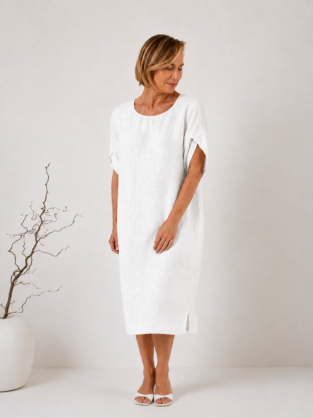 A woman modeling a loose-fitting white linen midi dress with short sleeves and a relaxed silhouette, standing in a minimalist studio setting with a white vase of branches on the left.