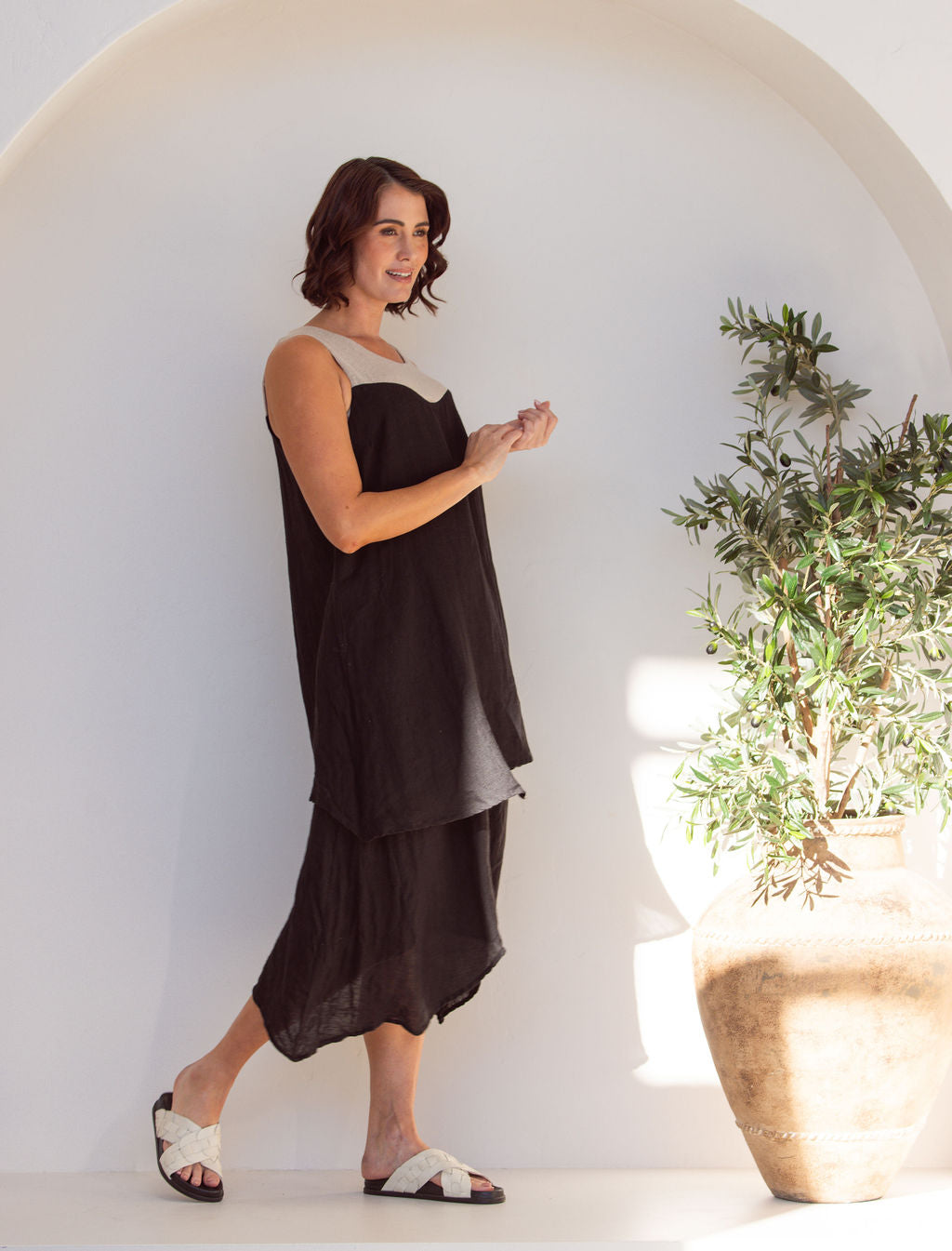Woman wearing a black and beige linen See Saw midi dress. Standing next to a plant in an arch minimal background.
