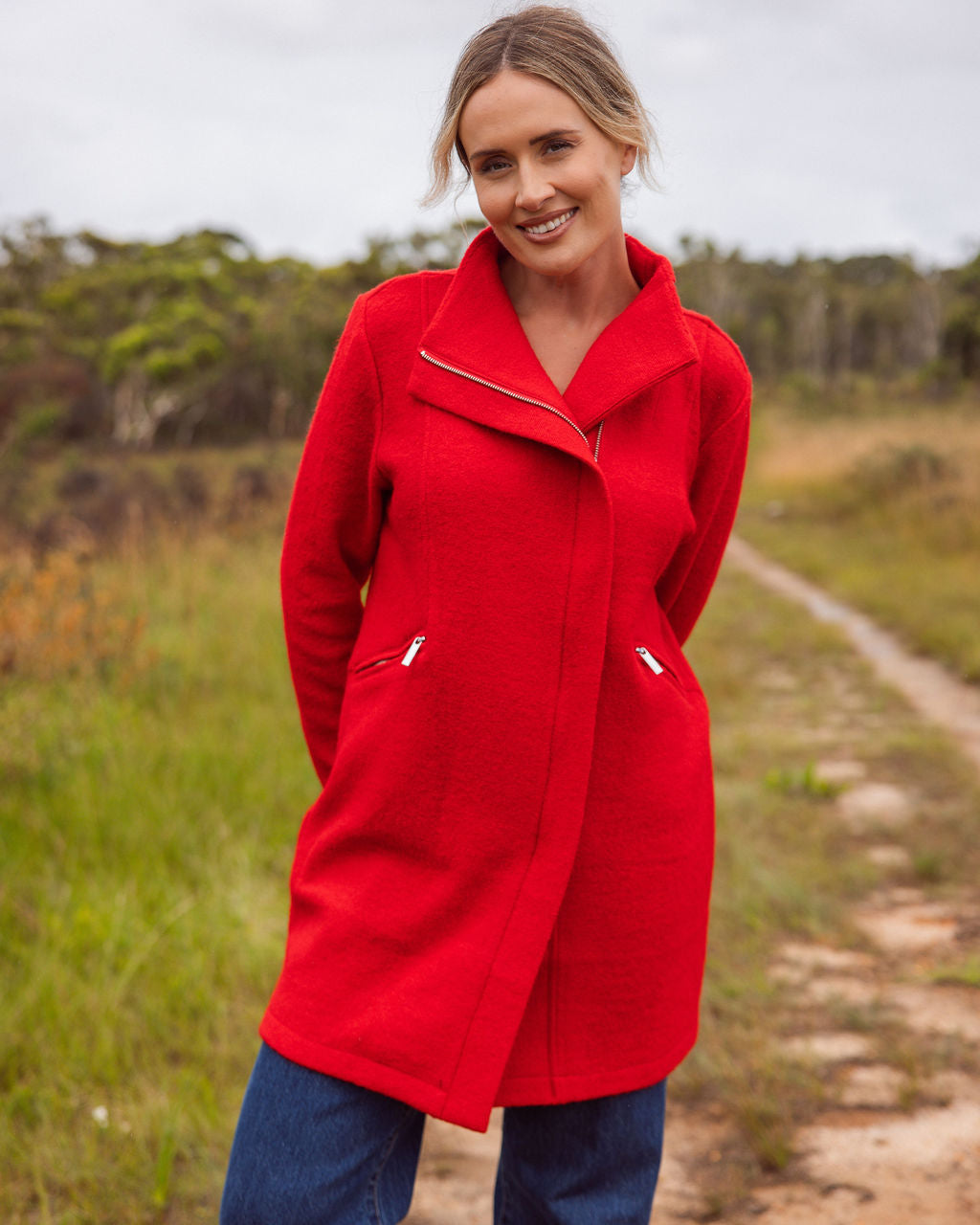 Woman wearing a See Saw boiled wool
red coat standing on a dirt path with trees in the background