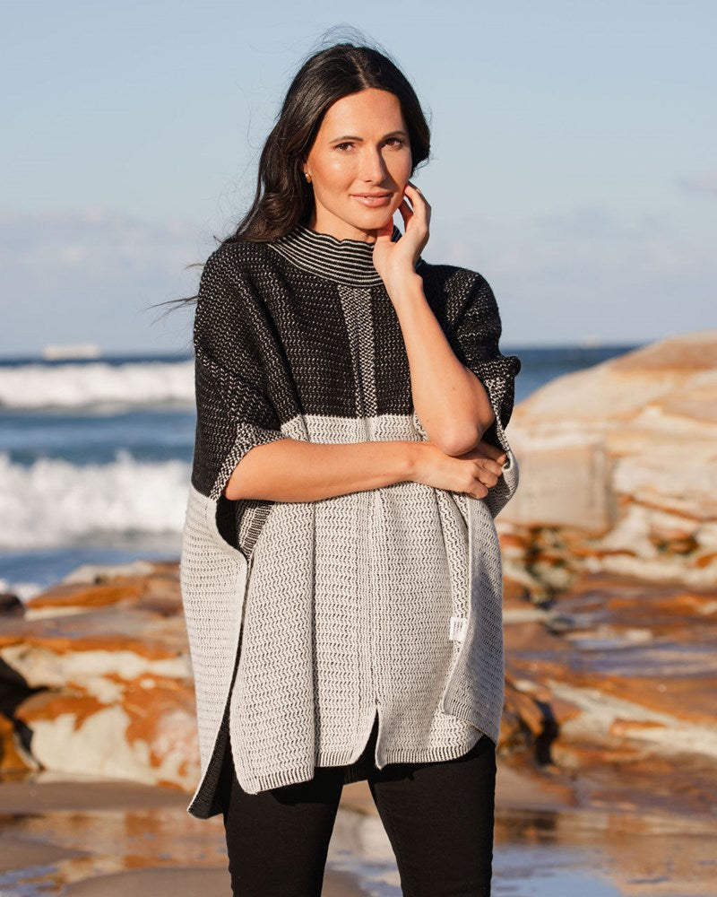 Woman wearing a See Saw wool blend black and grey patterned poncho on a beach with ocean and rocks in the background