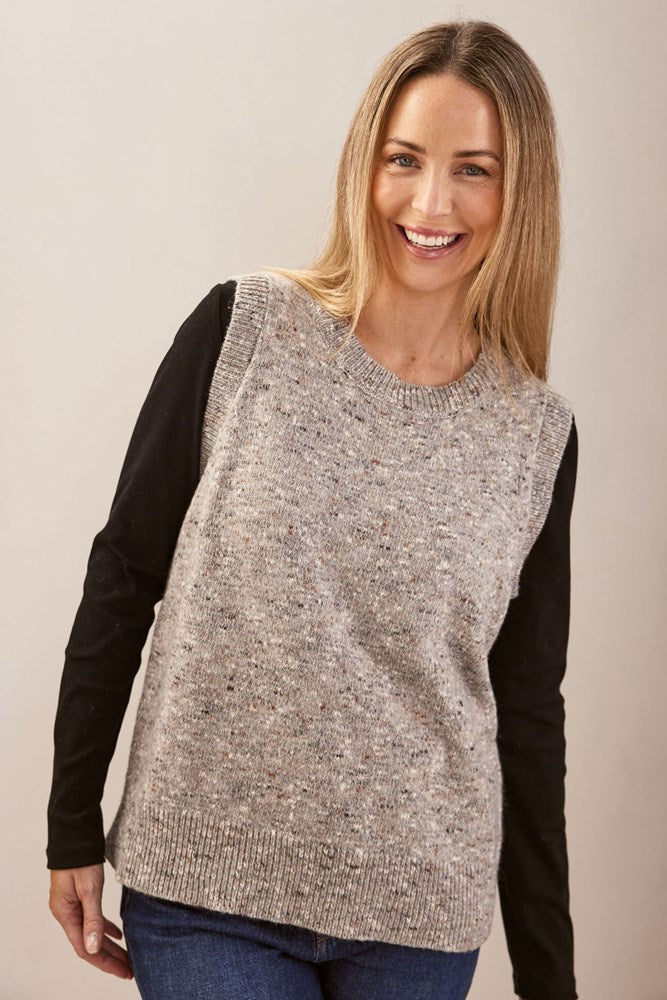 Woman wearing a beige knitted See Saw vest over a black long-sleeve shirt on a plain background