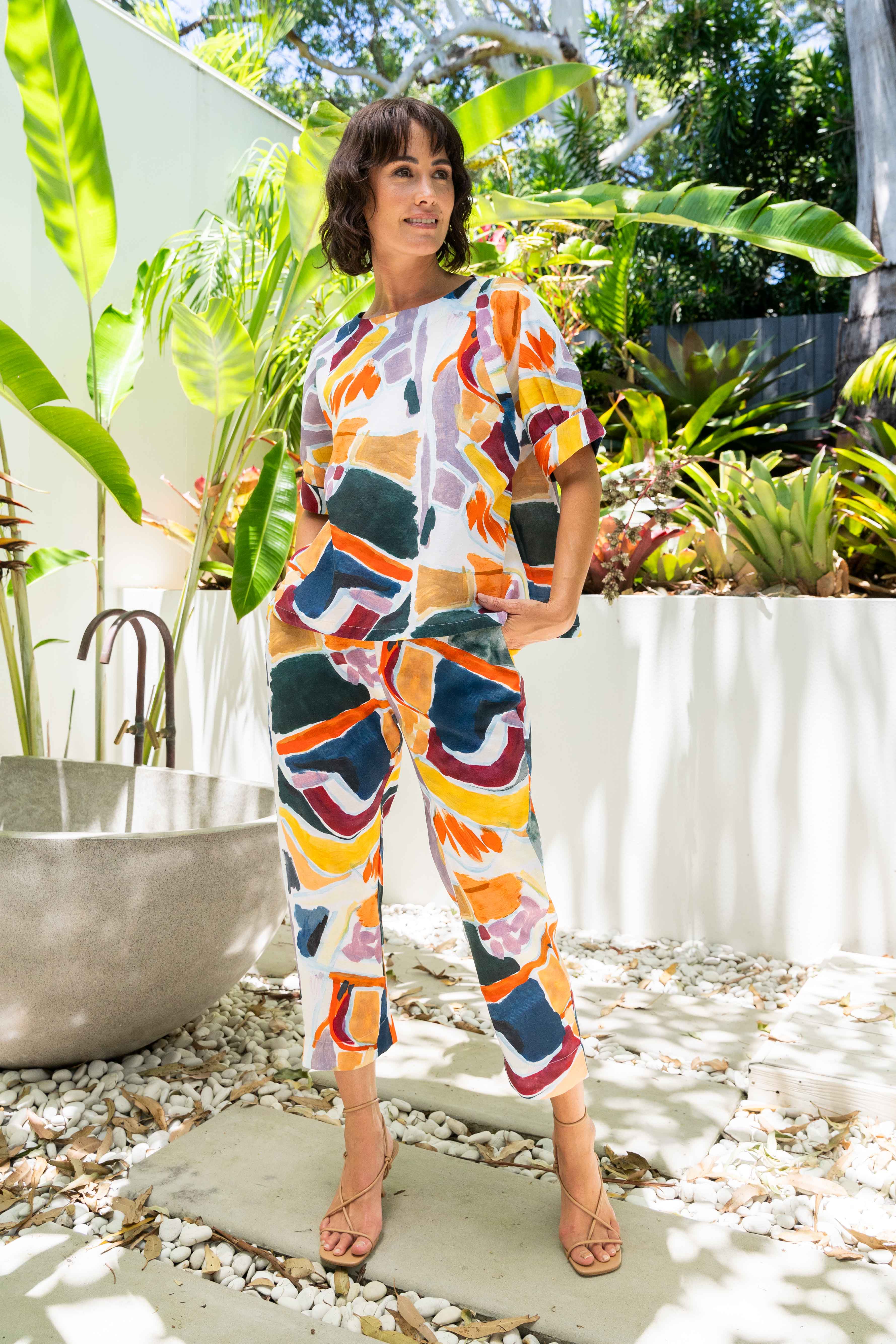 Woman in a colorful See Saw Linen pant set standing in a garden with plants and a chair.