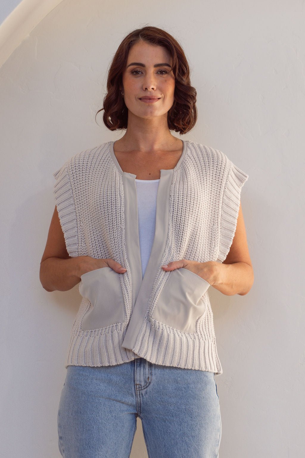Woman wearing a stone cotton See Saw knitted vest over a white shirt and blue jeans against a white wall.