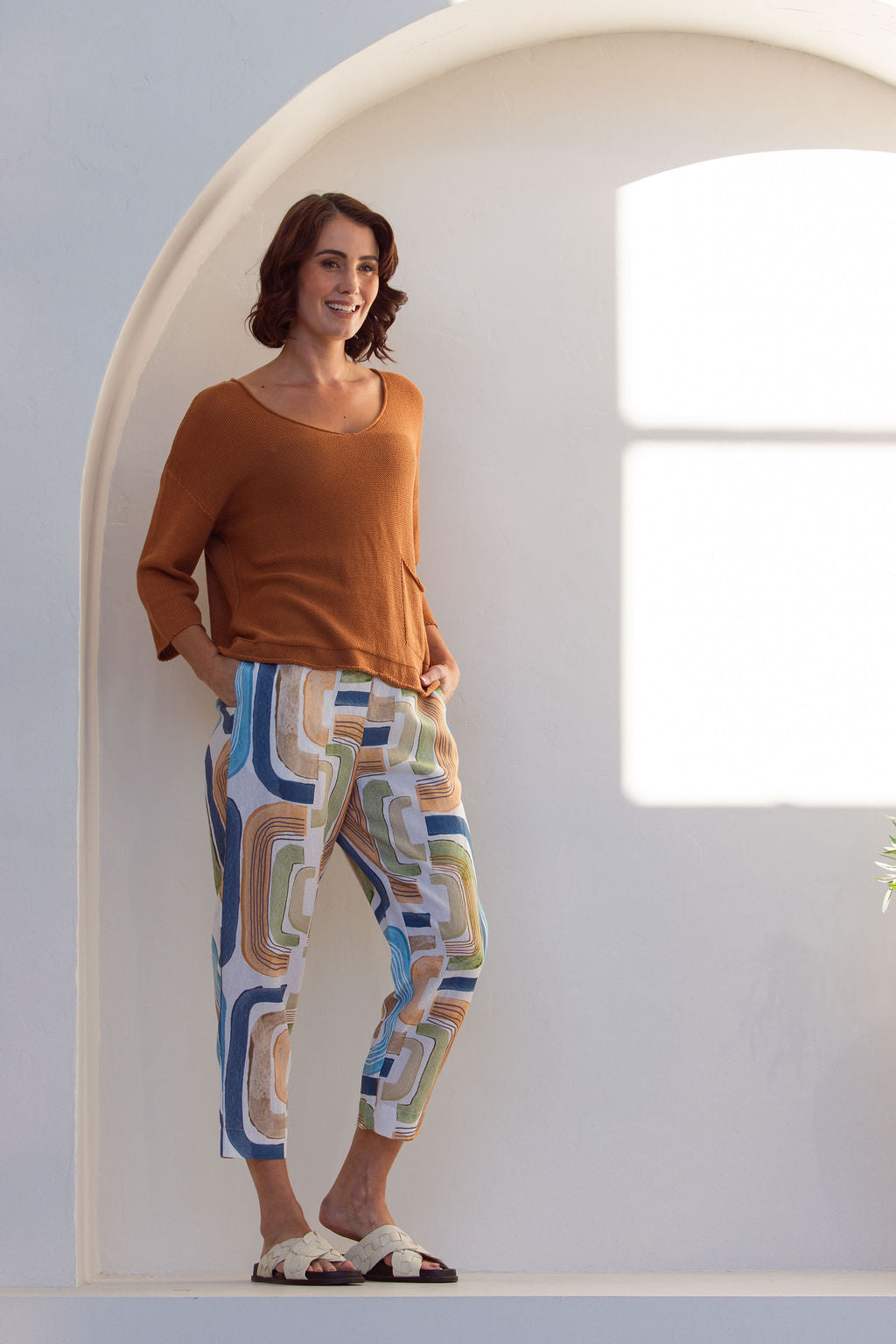 Woman wearing abstract See Saw seascape pant and rust top standing in a bright, sunlit room with white walls and a plant.