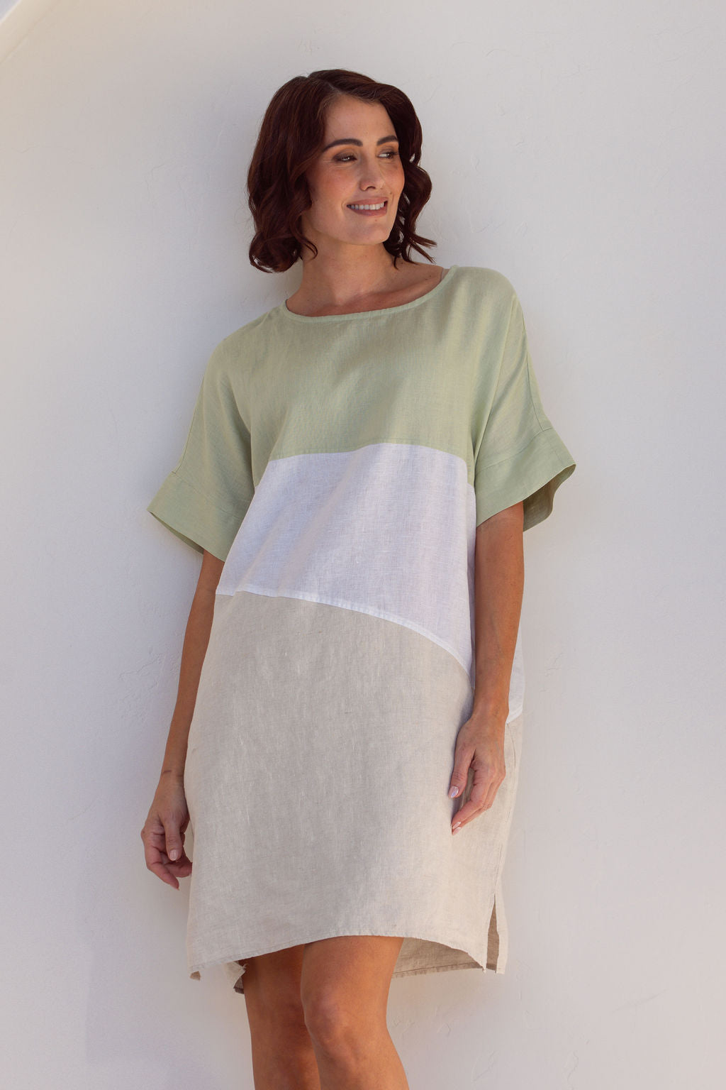 Woman wearing a Linen See Saw dress with a spliced pattern standing against a white wall with a plant to the side.