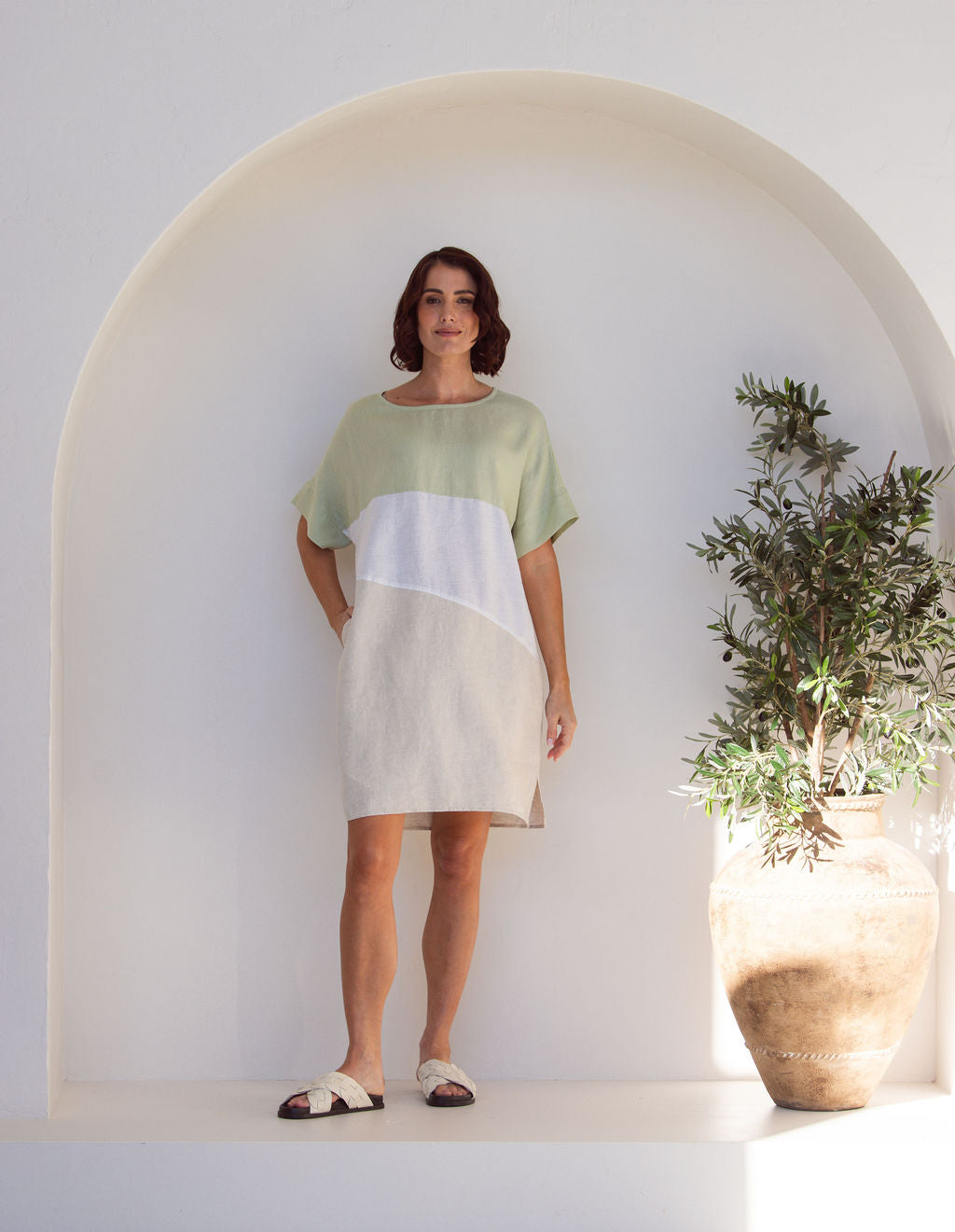 Woman wearing a Linen See Saw dress with a spliced pattern standing against a white wall with a plant to the side.