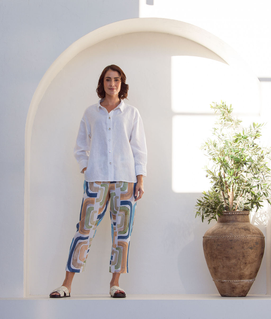 Woman wearing abstract See Saw seascape pant and white shirt standing in a bright, sunlit room with white walls and a plant.