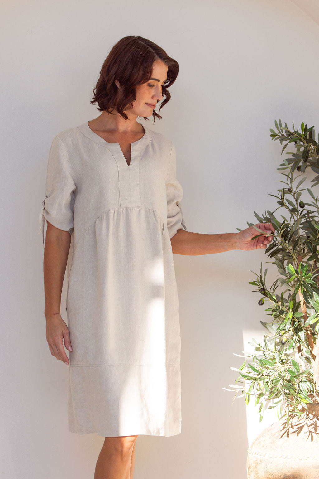 Woman in a Natural See Saw Dress with a gathered sleeve, standing inside an archway with a plant to the side.