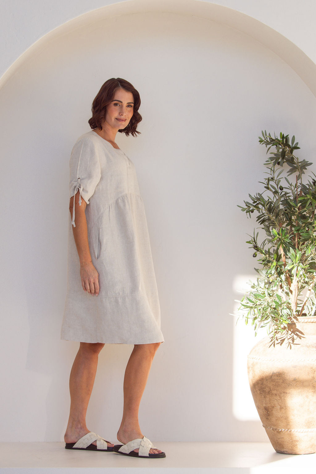 Woman in a Natural See Saw Dress with a gathered sleeve, standing inside an archway with a plant to the side.