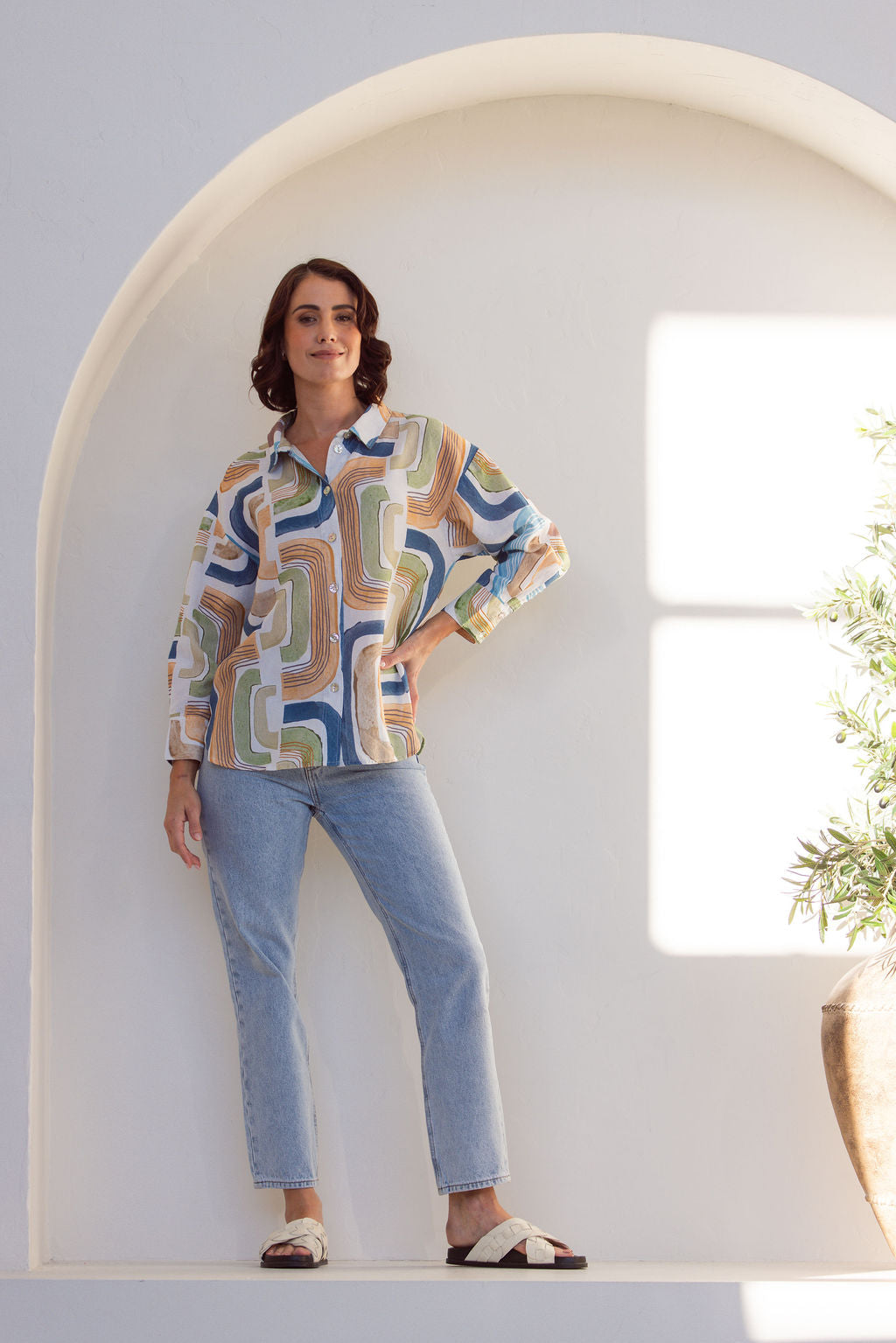 Woman wearing a long sleeve See Saw patterned linen button up shirt with jeans. Woman is standing next to a plant on a white background.