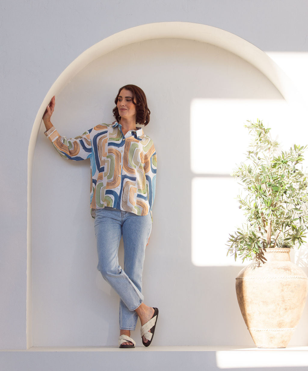 Woman wearing a long sleeve See Saw patterned linen button up shirt with jeans. Woman is standing next to a plant on a white background.
