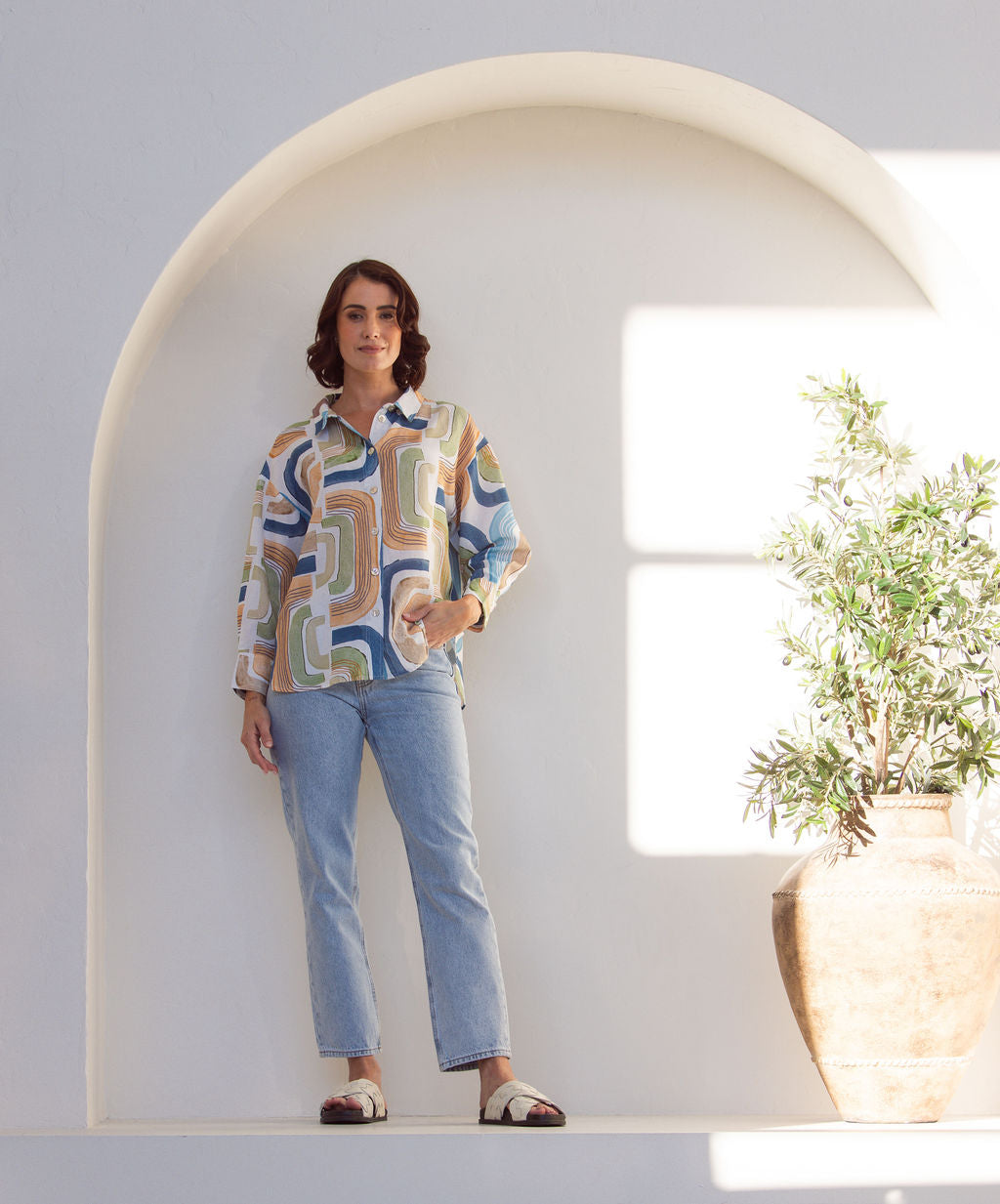 Woman wearing a long sleeve See Saw patterned linen button up shirt with jeans. Woman is standing next to a plant on a white background.