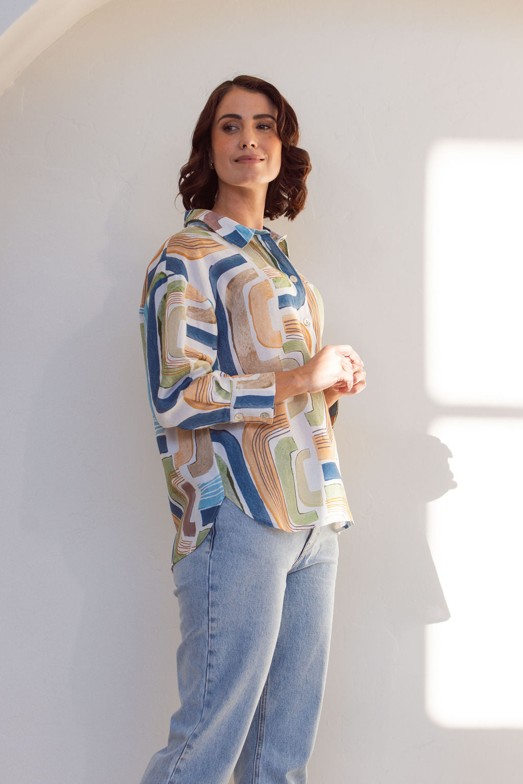 Woman wearing a long sleeve See Saw patterned linen button up shirt with jeans. Woman is standing in front of a white background.