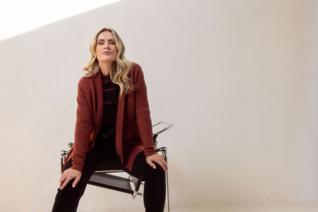 Woman wearing a brown See Saw cardigan standing next to a chair against a white background