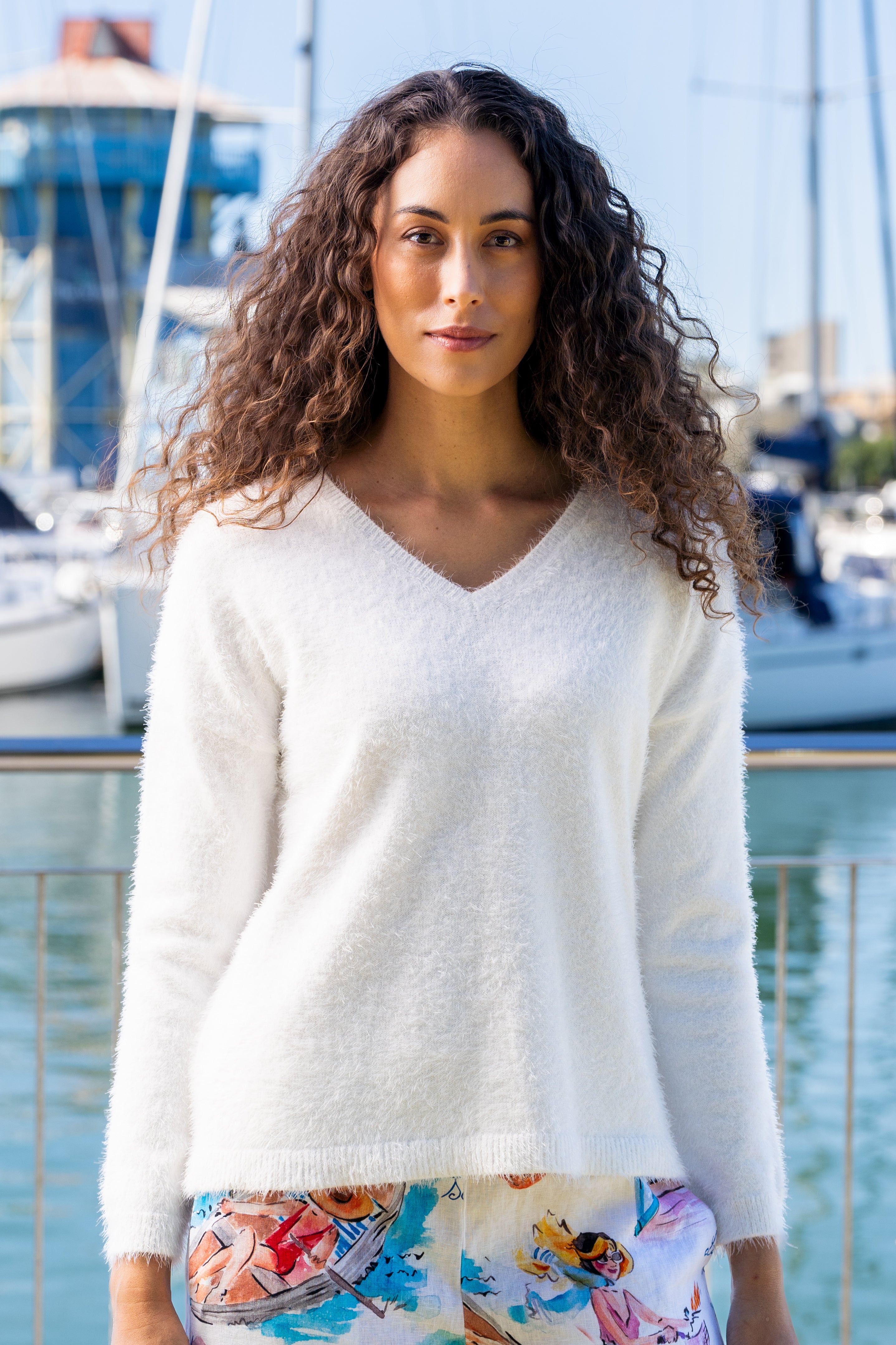 Woman wearing a white Kaleido textured sweater and colorful pants standing by a marina with boats in the background.