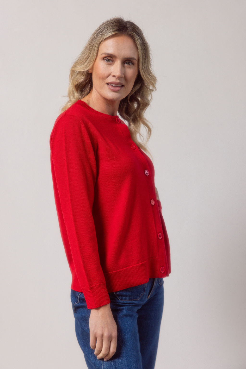 Woman wearing a merino wool See Saw red cardigan and blue jeans against a plain background