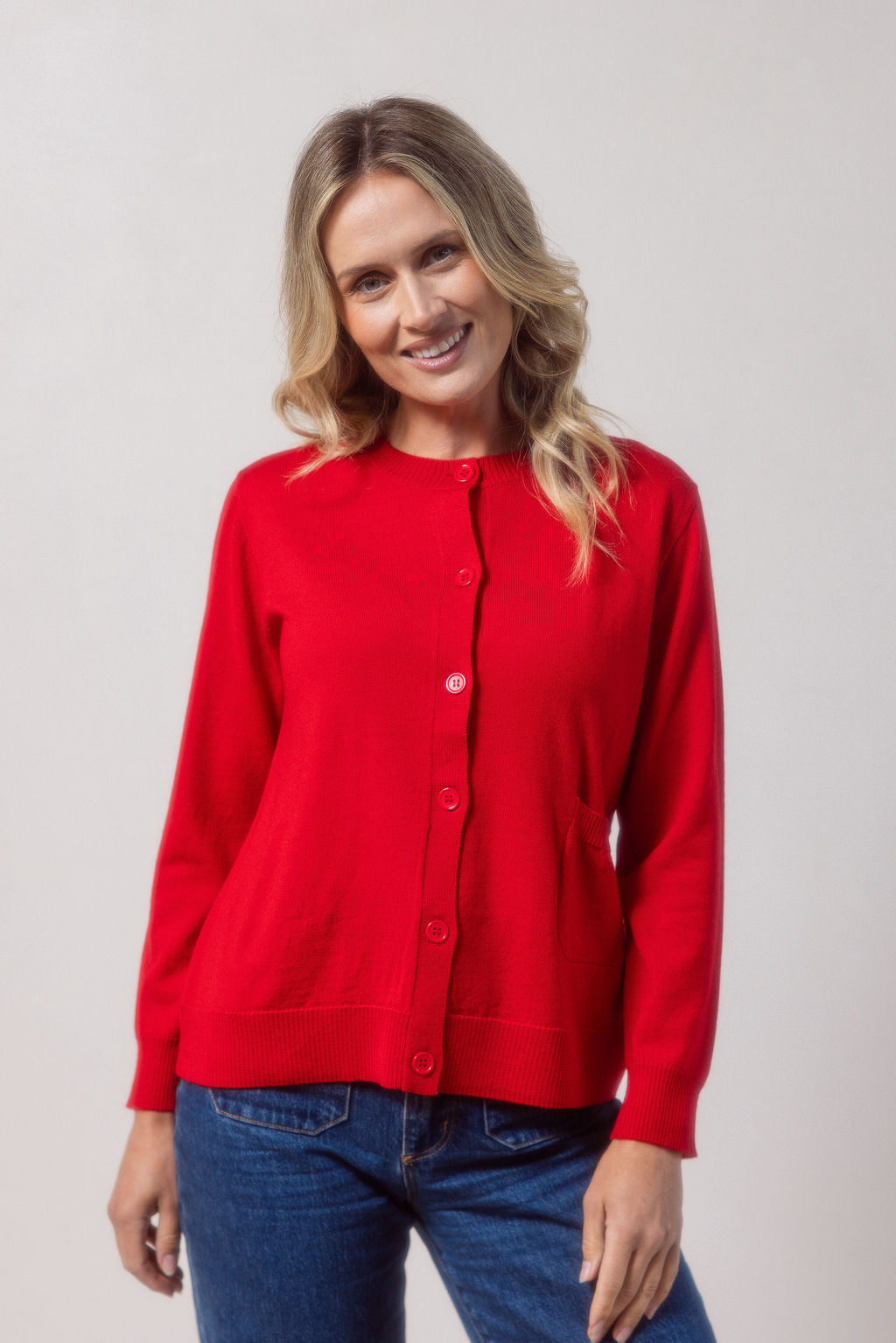 Woman wearing a merino wool See Saw red cardigan and blue jeans against a plain background