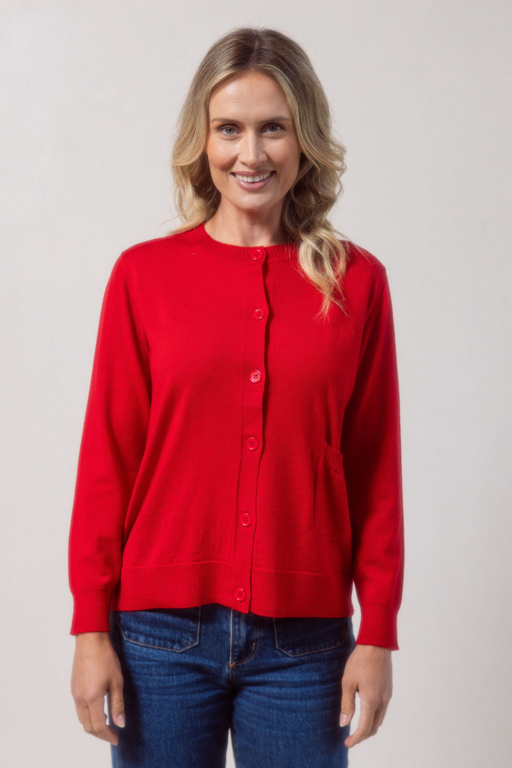 Woman wearing a merino wool See Saw red cardigan and blue jeans against a plain background