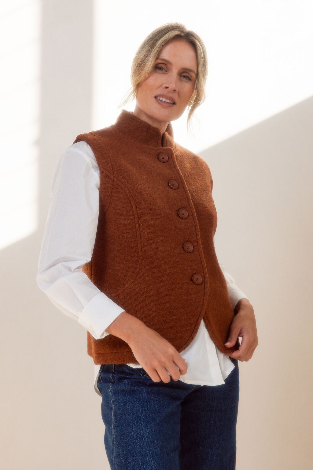 Woman wearing a brown vest over a white shirt and blue jeans against a neutral background