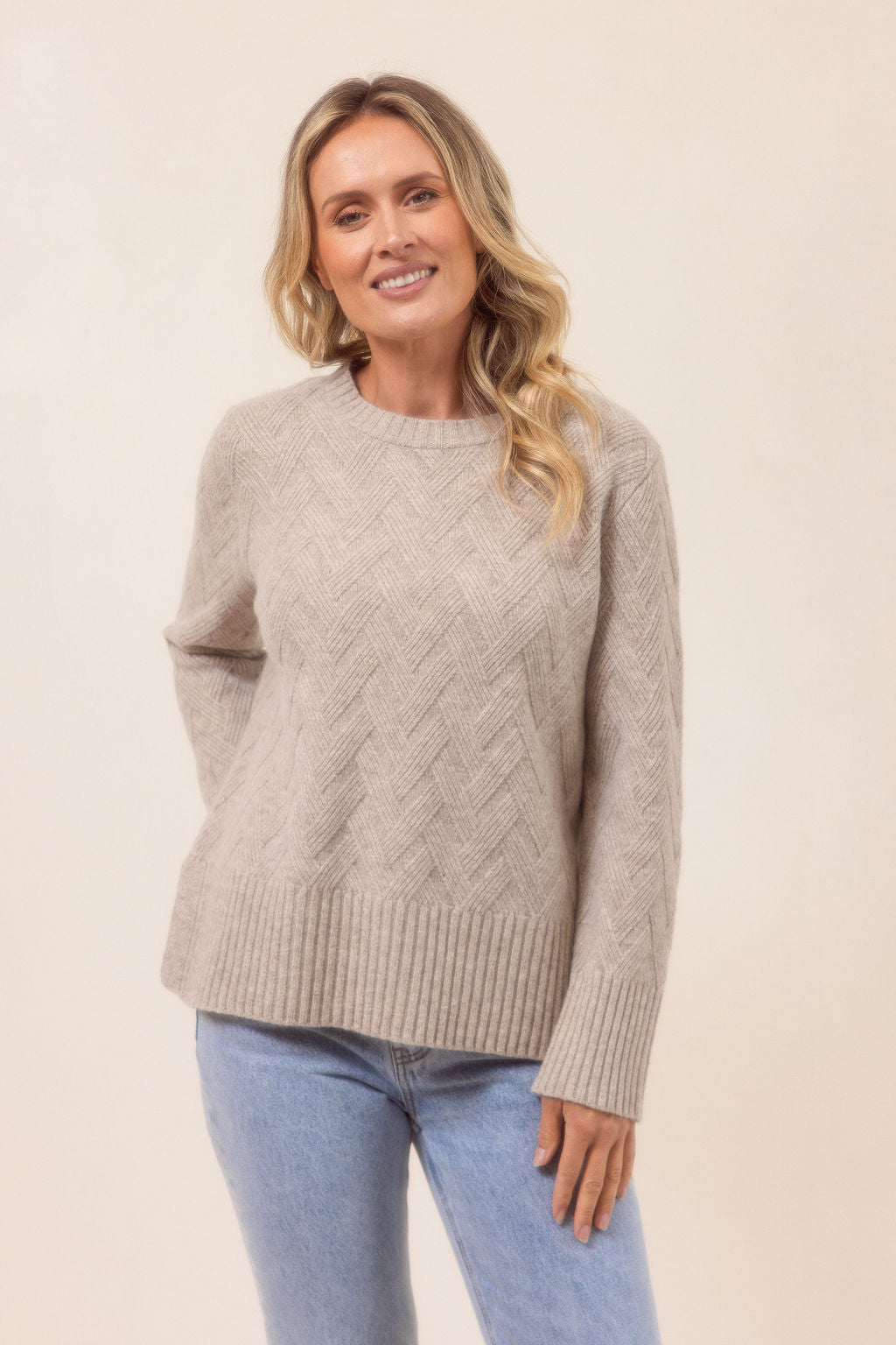 Woman wearing a beige See Saw cable knit sweater and blue jeans on a beige background