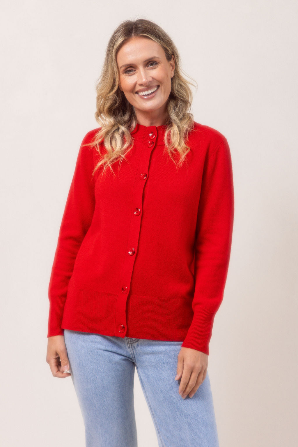 Woman wearing a red See Saw merino wool cardigan and light blue jeans against a plain background