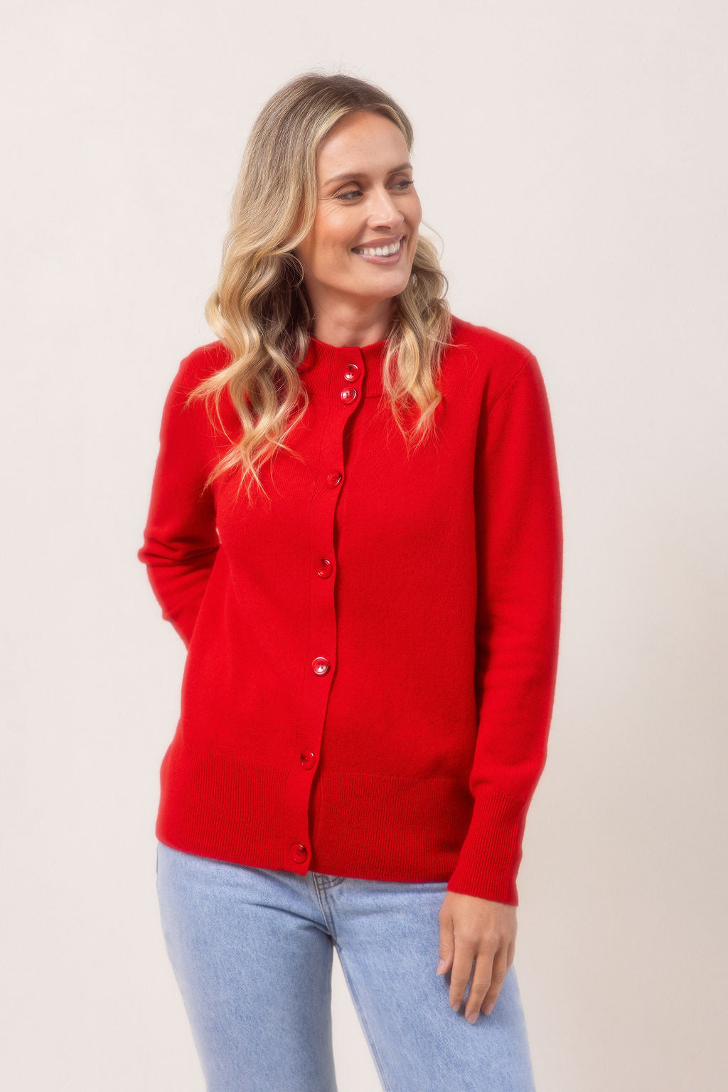 Woman wearing a red See Saw merino wool cardigan and light blue jeans against a plain background