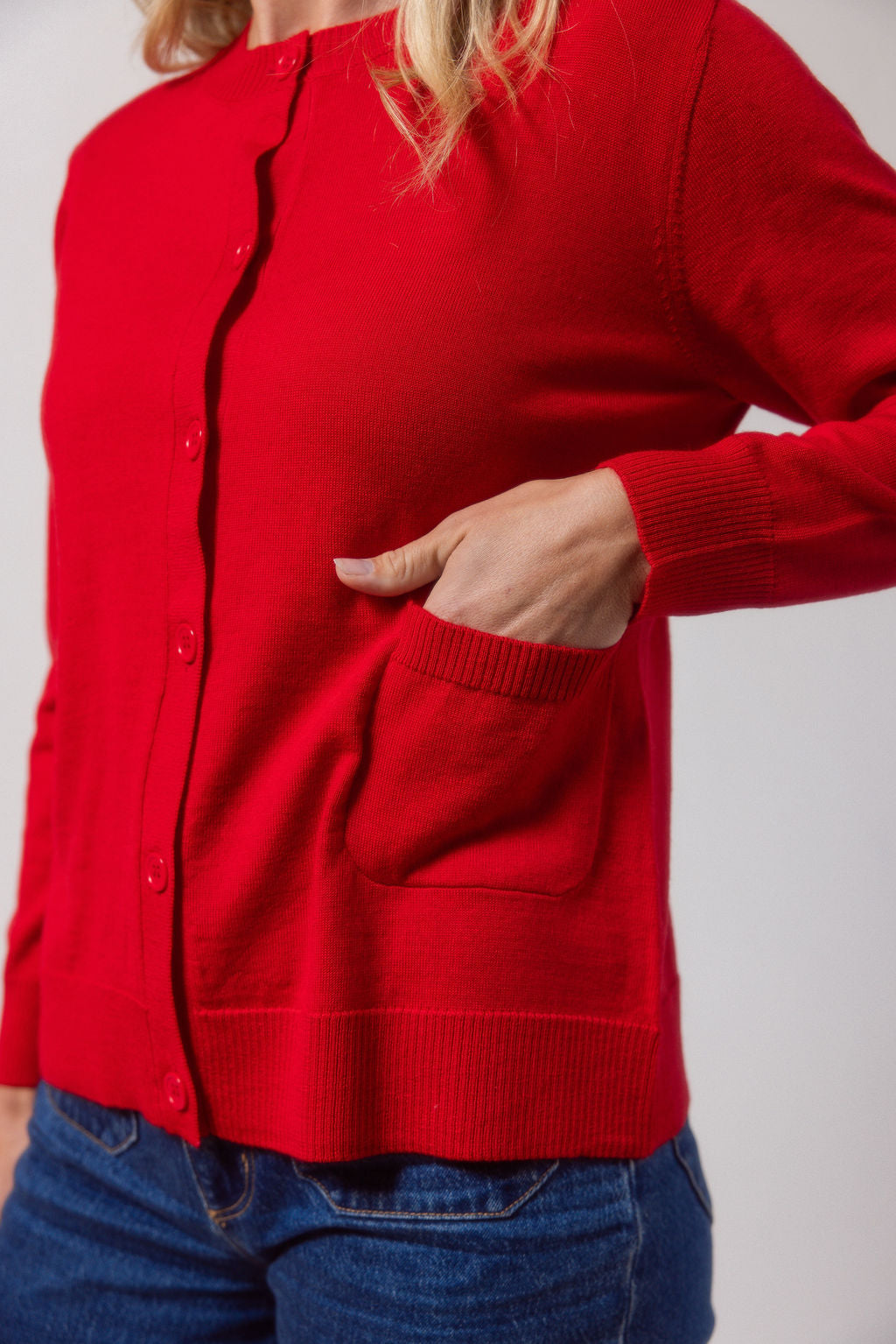 Woman wearing a red See Saw merino wool cardigan and light blue jeans against a plain background