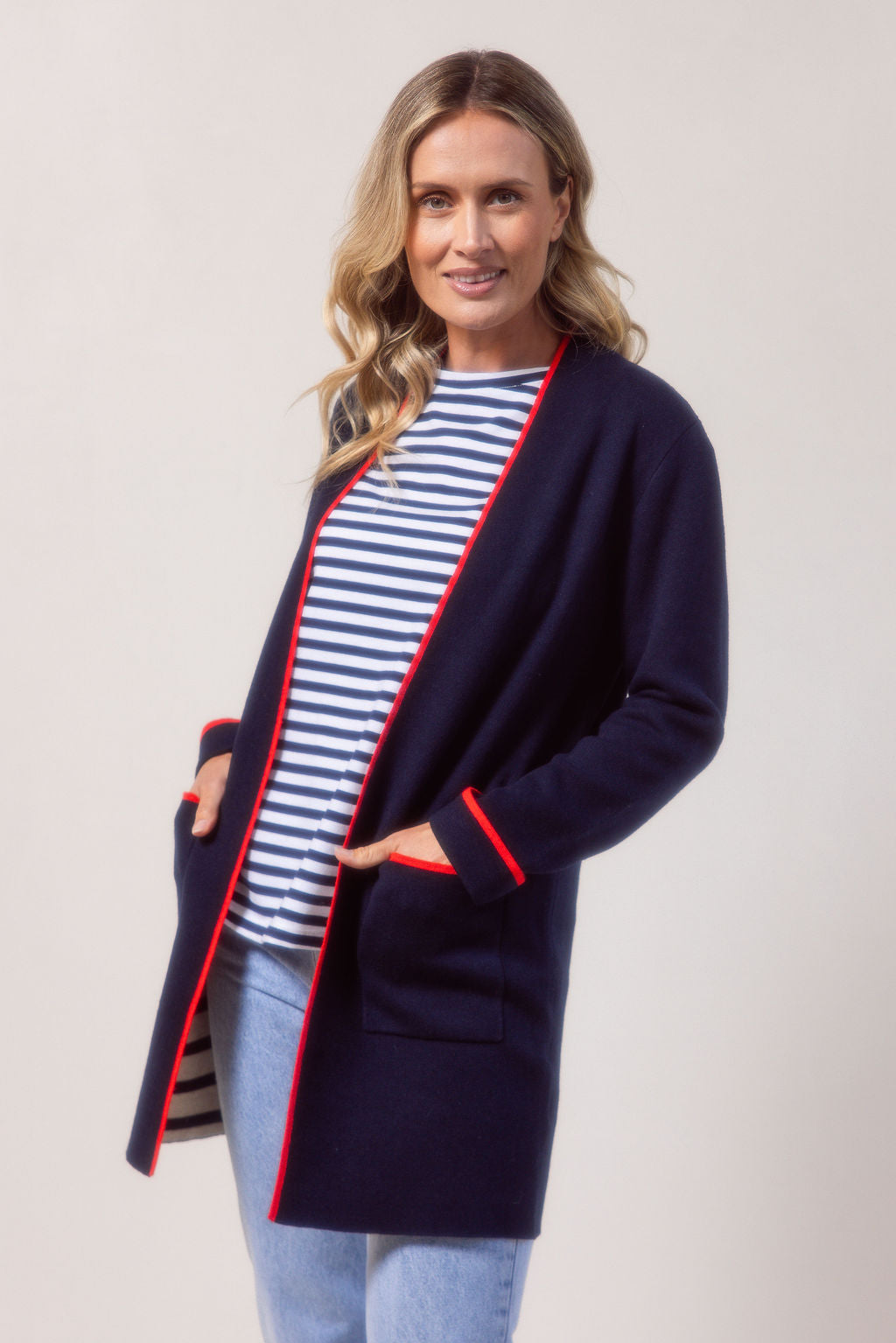 Woman wearing a See Saw navy cardigan with red trim, striped shirt, jeans, and white sneakers on a plain background