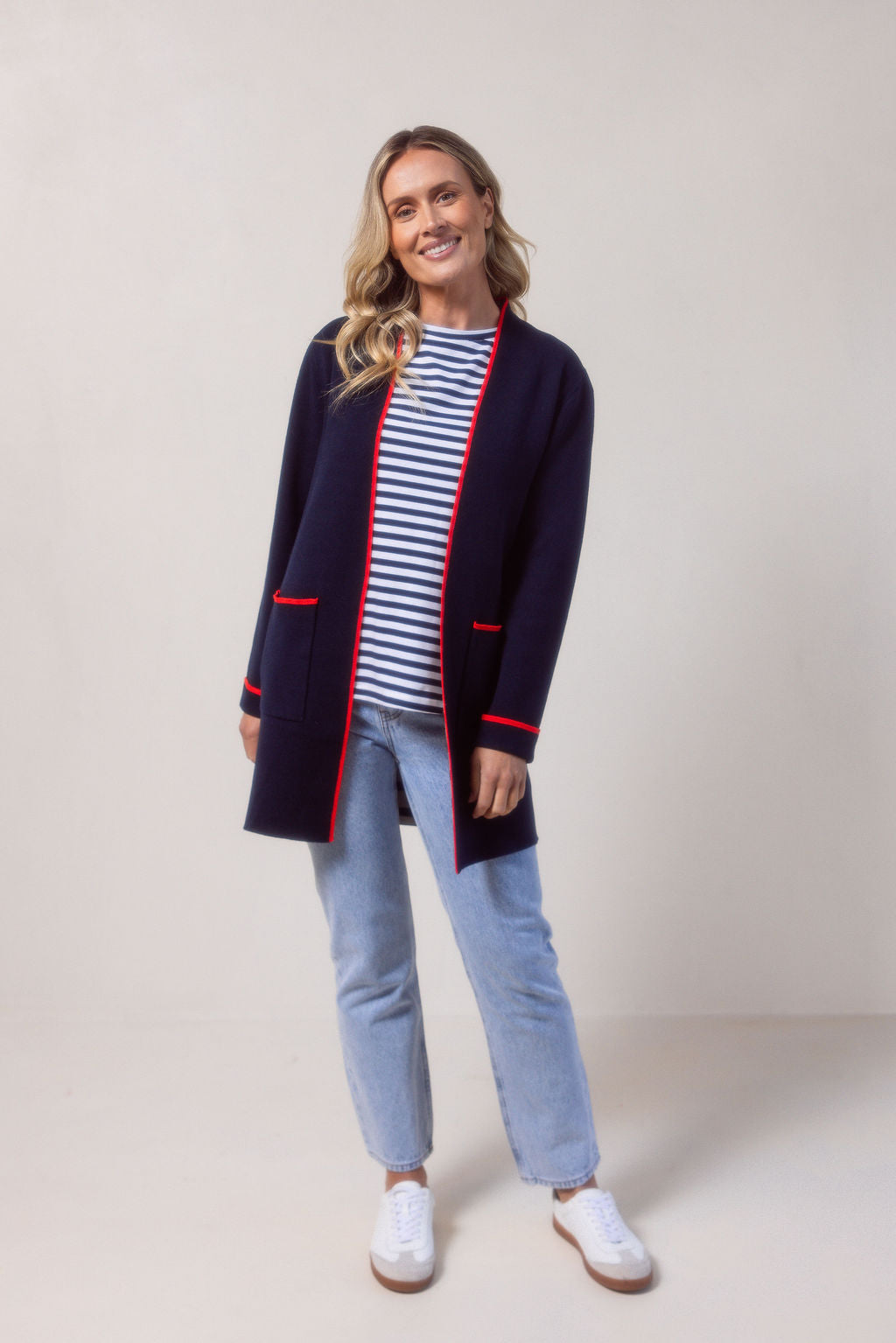 Woman wearing a See Saw navy cardigan with red trim, striped shirt, jeans, and white sneakers on a plain background