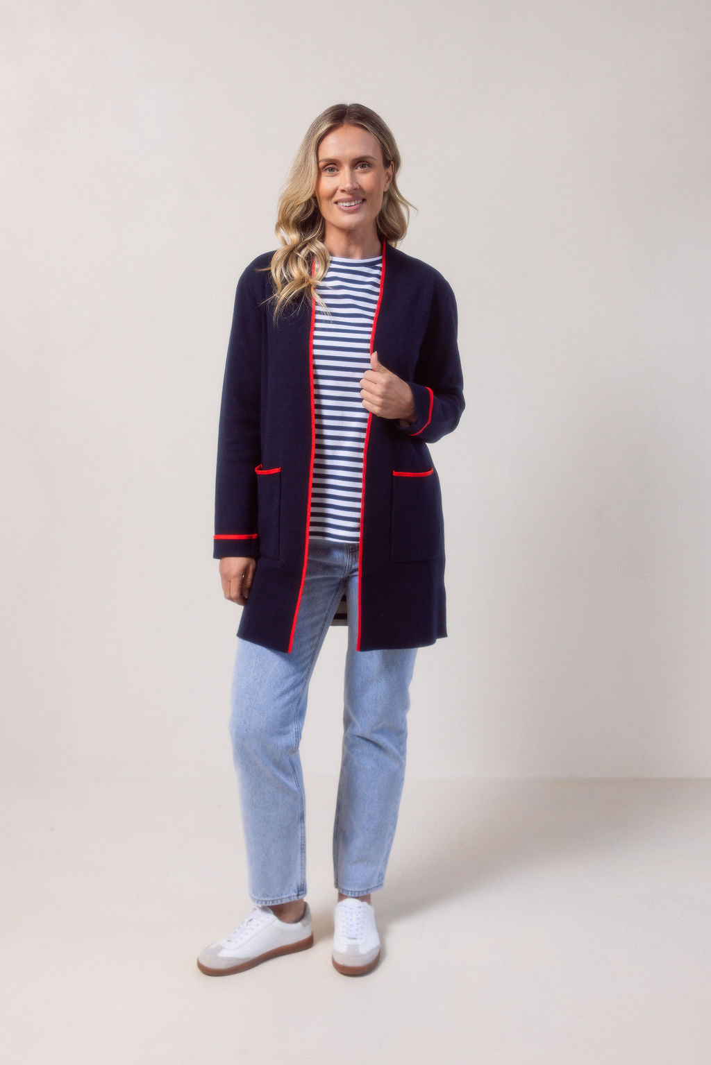 Woman wearing a See Saw navy cardigan with red trim, striped shirt, jeans, and white sneakers on a plain background