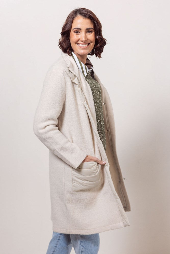 Woman wearing a cream boiled wool See Saw coat over a patterned sweater on a plain background