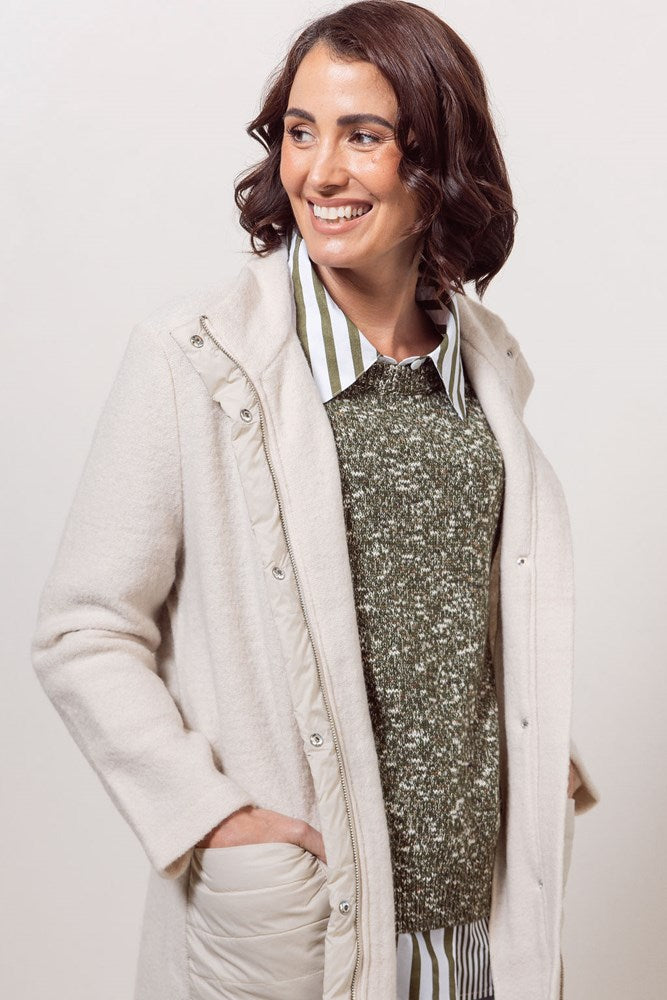 Woman wearing a cream boiled wool See Saw coat over a patterned sweater on a plain background