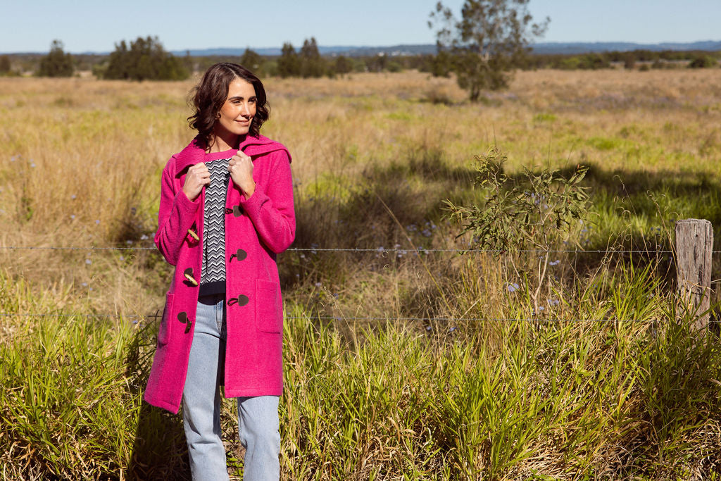 Woman wearing a pink See Saw Duffle coat in boiled wool standing in a field with trees and sky in the background