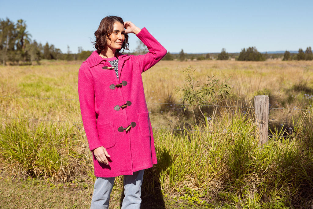 Woman wearing a pink See Saw Duffle coat in boiled wool standing in a field with trees and sky in the background