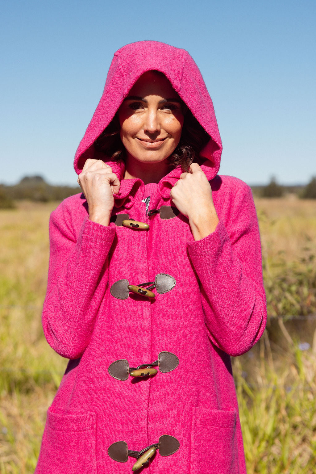 Woman wearing a pink See Saw Duffle coat in boiled wool standing in a field with trees and sky in the background