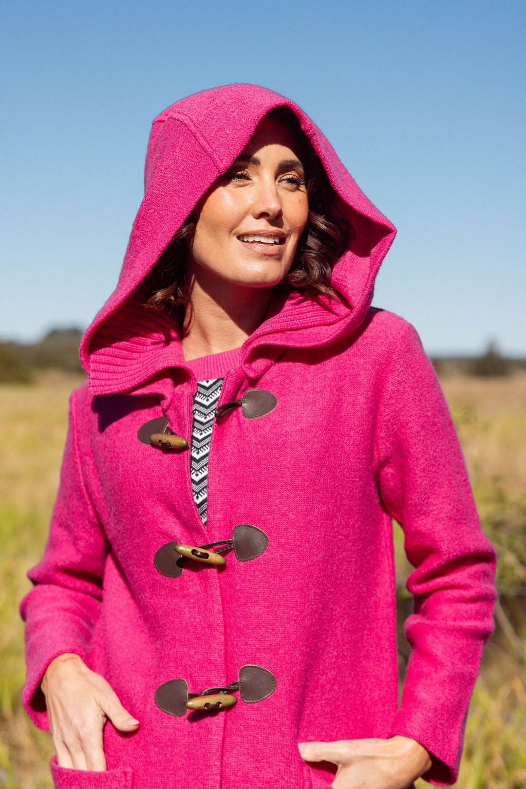 Woman wearing a pink See Saw Duffle coat in boiled wool standing in a field with trees and sky in the background