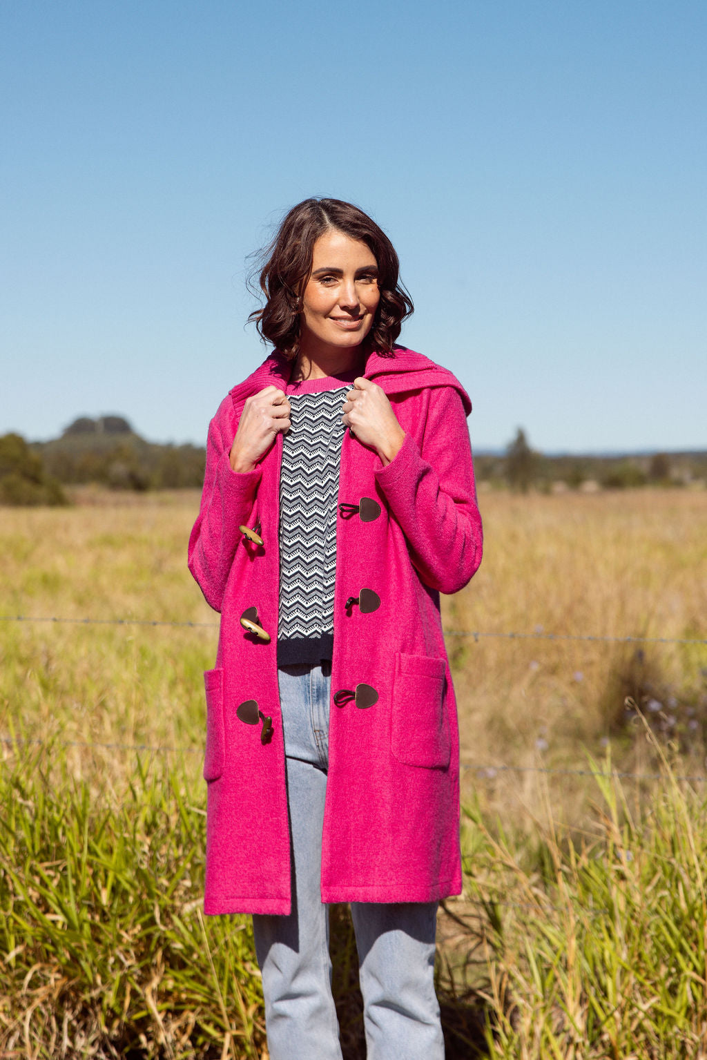 Woman wearing a pink See Saw Duffle coat in boiled wool standing in a field with trees and sky in the background