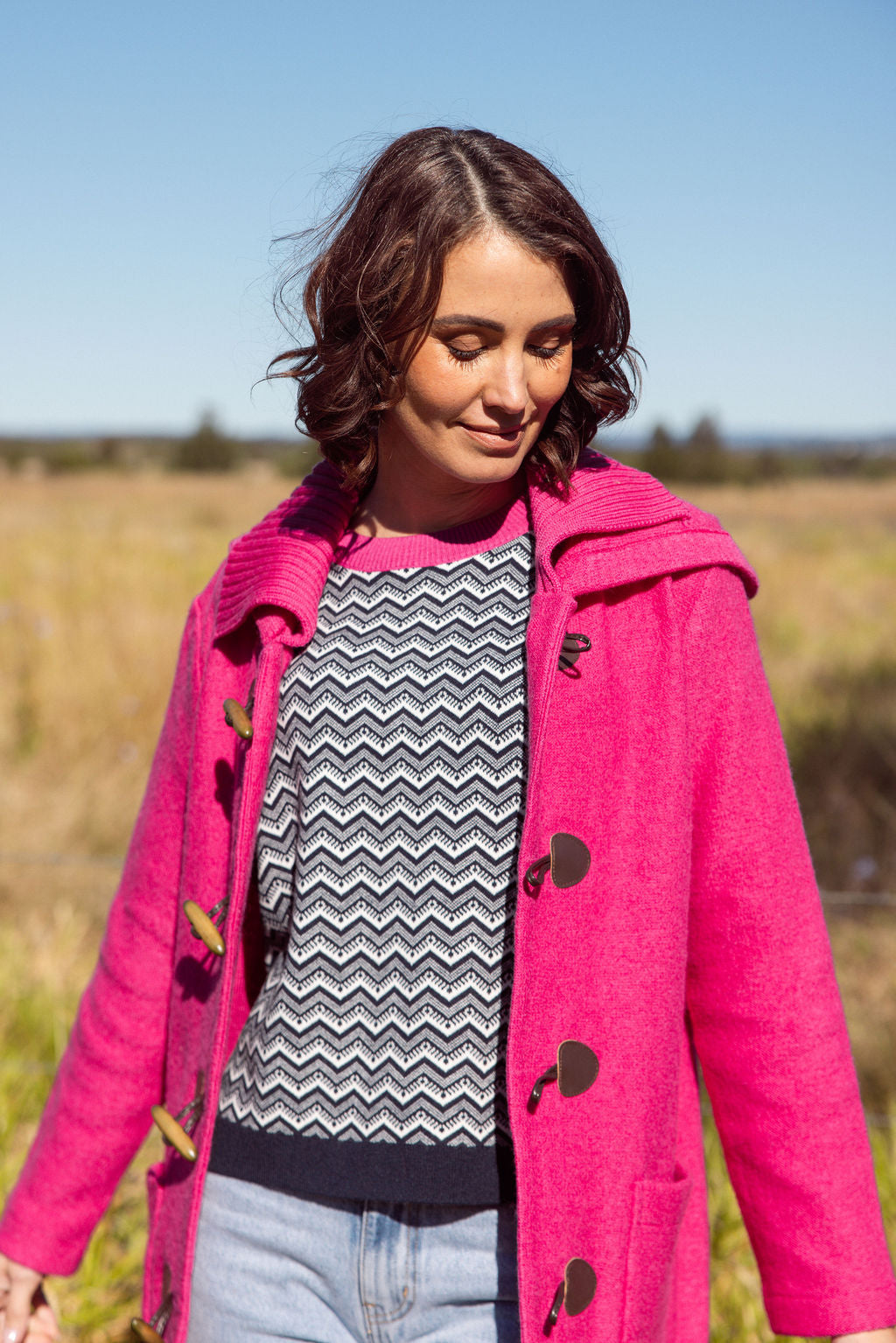 Woman wearing a pink See Saw Duffle coat in boiled wool standing in a field with trees and sky in the background