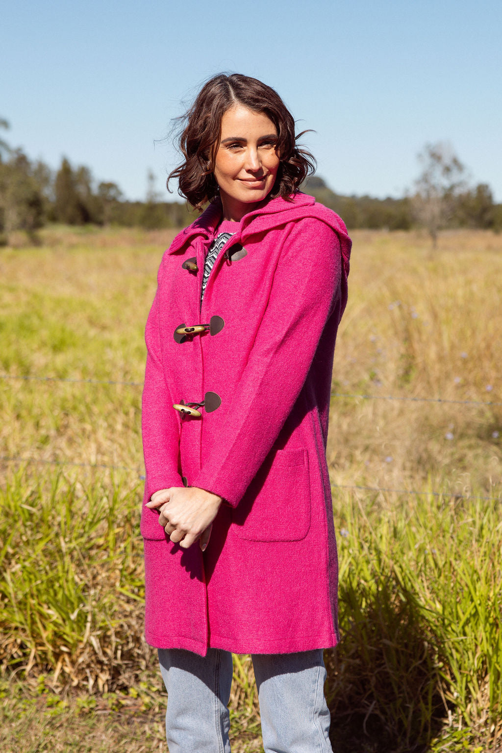 Woman wearing a pink See Saw Duffle coat in boiled wool standing in a field with trees and sky in the background