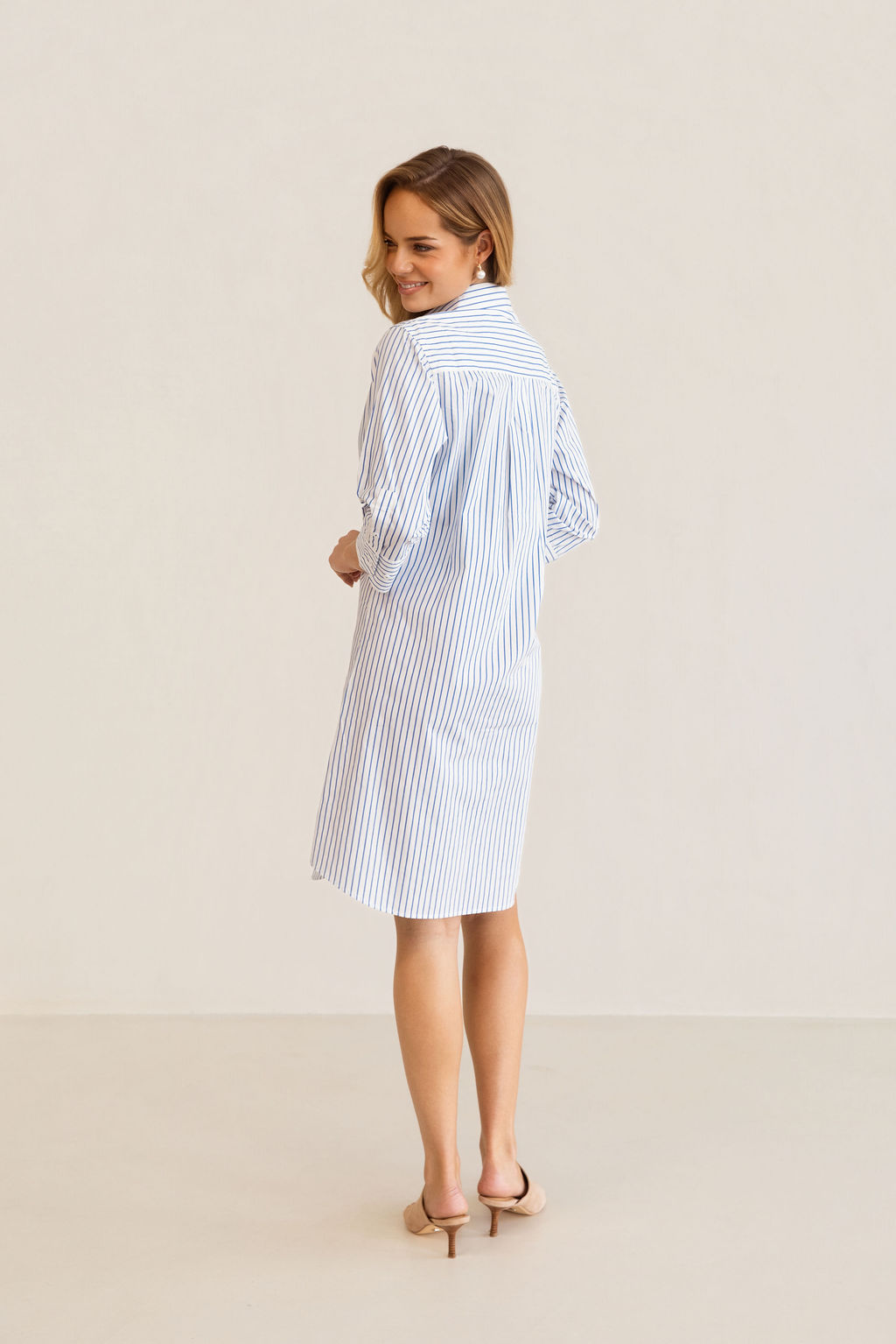 A woman modeling a blue and white Kaleido cotton vertically striped button-down shirt dress with three-quarter sleeves, standing against a plain off-white studio background.