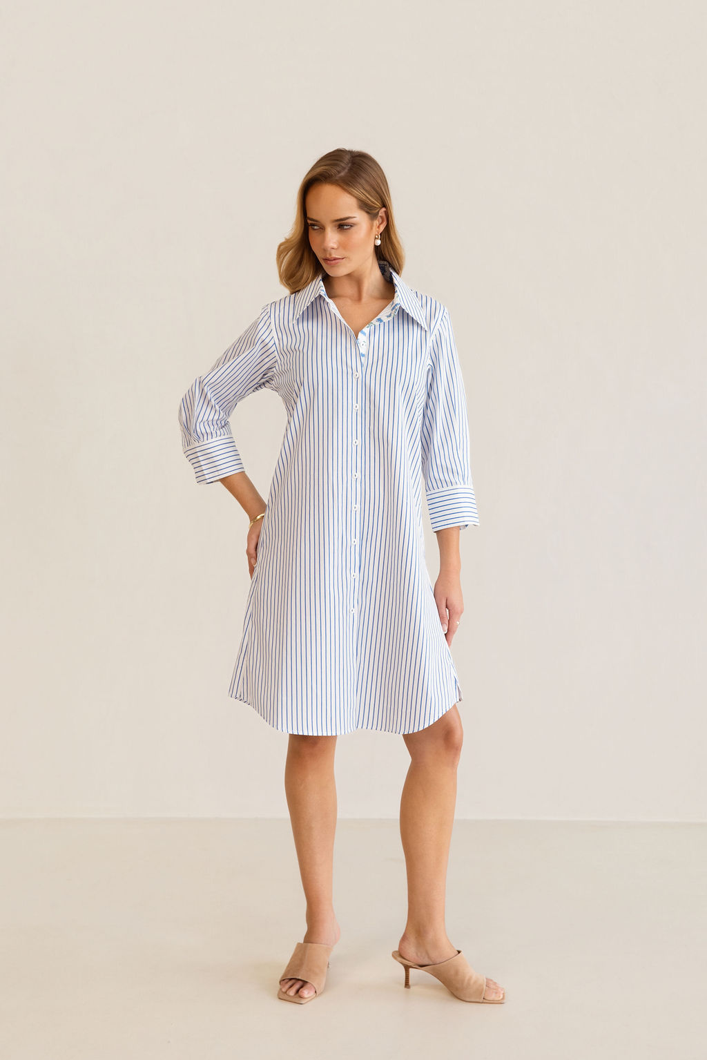 A woman modeling a blue and white Kaleido cotton vertically striped button-down shirt dress with three-quarter sleeves, standing against a plain off-white studio background.