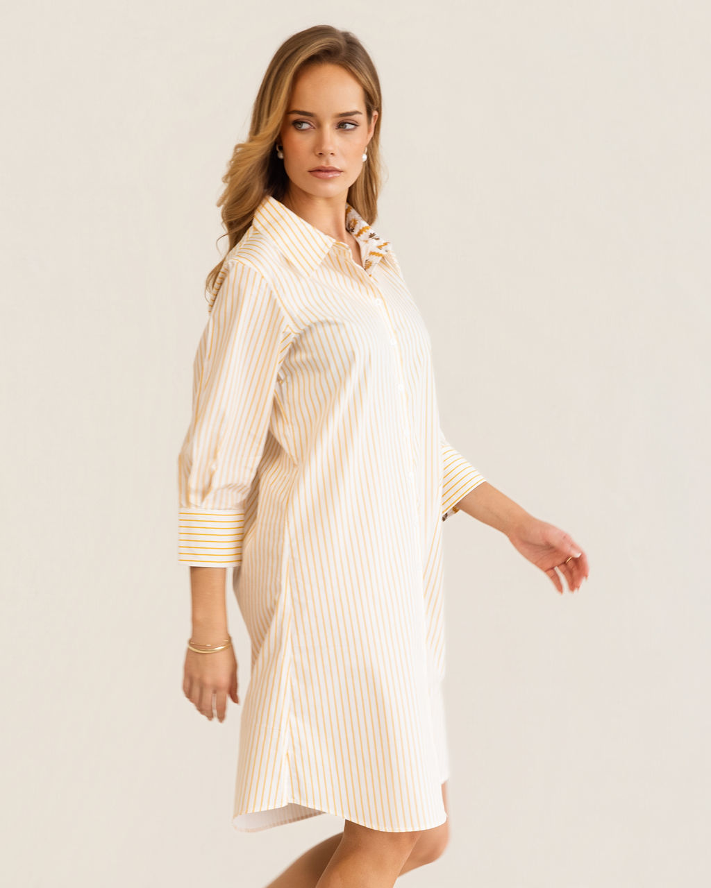 A woman modeling a yellow and white Kaleido cotton vertically striped button-down shirt dress with three-quarter sleeves, standing against a plain off-white studio background.