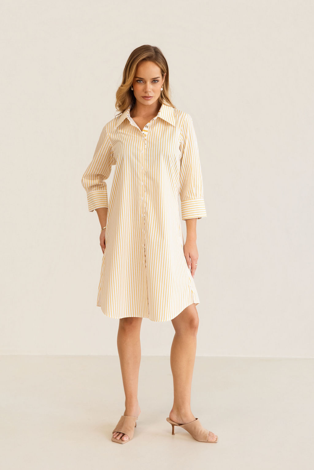 A woman modeling a yellow and white Kaleido cotton vertically striped button-down shirt dress with three-quarter sleeves, standing against a plain off-white studio background.