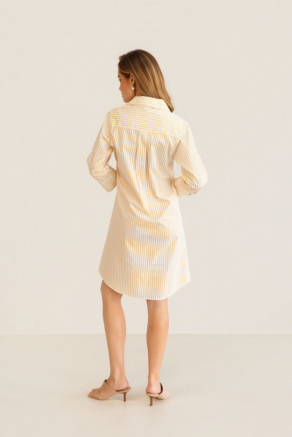 A woman modeling a yellow and white Kaleido cotton vertically striped button-down shirt dress with three-quarter sleeves, standing against a plain off-white studio background.